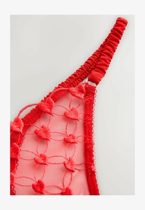 Red bralette made of mesh and lace with heart-shaped embroidery, featuring a ruffled strap and scalloped edges for texture and detail.