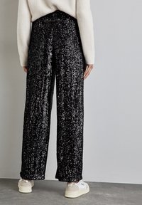 Czarne szerokie spodnie z cekinami, z wysokim stanem i teksturowaną powierzchnią, zestawione z białymi sneakersami. Główne akcenty to błyszczące ozdoby.