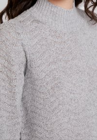 Pull en tricot gris clair avec un col montant, présentant un motif texturé en forme de vague. Le matériau semble doux et légèrement transparent.