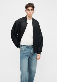 Jaunas vyras dėvi juodą bombonešio striukę ant balto marškinėlių ir mėlynų džinsų, stovintis prieš paprastą šviesiai pilką foną.