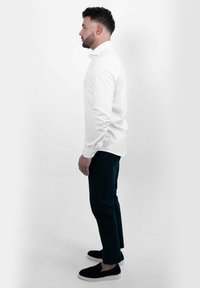 Witte overhemden met knopen, donkere broek en zwarte instappers. Het shirt heeft lange mouwen en een standaard kraag, gezien vanuit een zijprofiel.