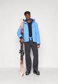 Lyžařský outfit složený z lehké modré izolační bundy, černého základního oblečení, tmavých kalhot a černých lyžařských bot, držící dřevěné lyže s vázáním.