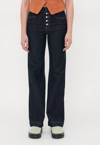 Mørkeblå høyt midjete jeans med rett passform, som har fem metallknapper, kontrastsøm og sidelommer. Båret med grønne sko.