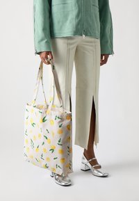Person wearing cream pants with front slits, silver block-heeled shoes, and a green jacket, holding a tote bag with lemon print.