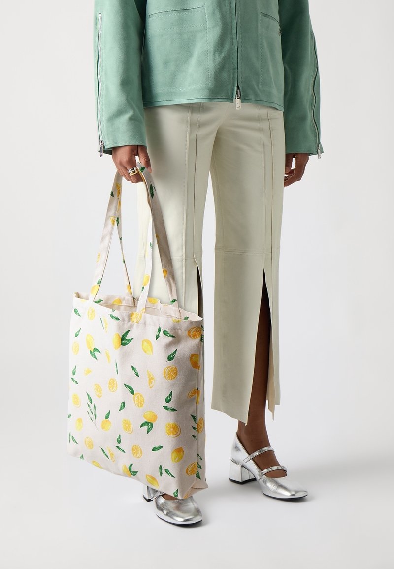 Person wearing cream pants with front slits, silver block-heeled shoes, and a green jacket, holding a tote bag with lemon print.