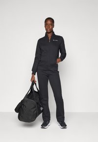 Veste zippée noire et pantalon assorti en tissu doux et texturé, accompagnés d'un grand sac de sport noir et de baskets noires.