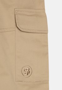 Pantalons cargo beiges avec une grande poche latérale, des coutures discrètes et un logo brodé circulaire. Fabriqués en tissu durable.
