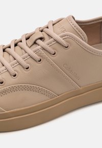 Beżowe skórzane sneakersy Calvin Klein z dopasowanymi sznurówkami, detalami przeszycia i płaską gumową podeszwą na białym tle.