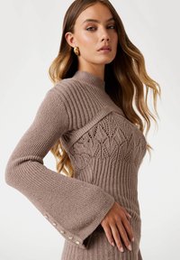 Frau in einem taupefarbenen Strickpullover mit kompliziertem Spitzenmuster auf der Brust, gerippten Ärmeln mit ausgestellten Bündchen und goldenen Knopfdetails.