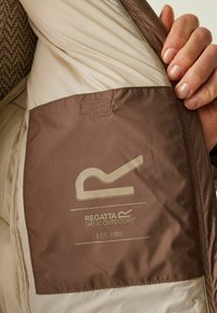 Mano sosteniendo una chaqueta beige abierta que revela un bolsillo interior marrón con el logo "Regatta Great Outdoors Est. 1985" impreso en su interior.