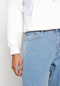 Hvit hettegenser med lavendel detaljer og kengurulomme, satt sammen med lys blå denimjeans med gullkomponenter og en myk tekstur.