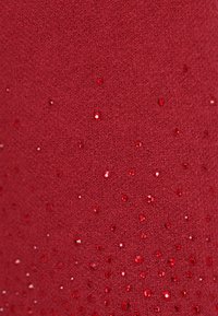 Červený textil s texturovaným povrchem ozdobený roztroušenými malými, lesklými červenými flitry, které vytvářejí jemný třpytivý efekt.