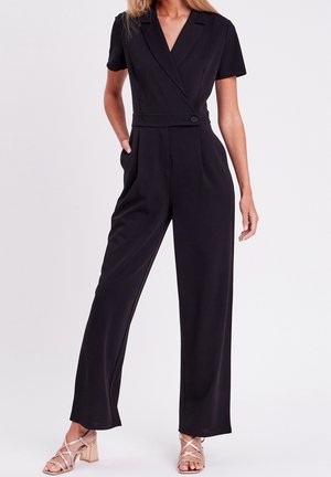 Femme portant une combinaison noire à manches courtes, jambes larges, avec une taille boutonnée et un col ouvert, associée à des sandales beiges à talons blocs.