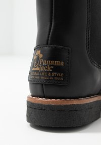 Botină din piele neagră, cu un toc din cauciuc brun și margine cusută. Include o etichetă cu logo-uri aurii cu numele brandului și țara de origine.