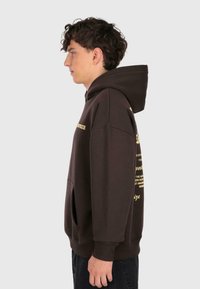 Un jeune homme avec des cheveux bouclés portant un sweat à capuche marron foncé avec du texte jaune, se tenant de profil devant un fond clair.