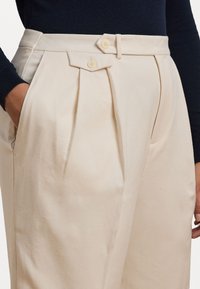 Pantaloni beige plissettati con vita alta, dotati di due bottoni e tasche laterali. Tessuto morbido con una finitura liscia.