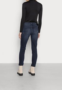 Tmavé džíny s úzkým střihem a pěticí kapsami, s polovičním pasem a jemným prošíváním. Kombinováno s béžovými kotníkovými botami.