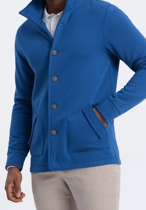 Homme portant une veste boutonnée bleu vif avec deux poches avant sur une chemise blanche et un pantalon beige, main dans la poche.