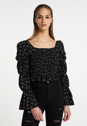 Junge Frau mit langen Haaren, die eine schwarze geraffte Bluse mit weißen Smiley-Gesichtern und zerrissene schwarze Jeans vor weißem Hintergrund trägt.