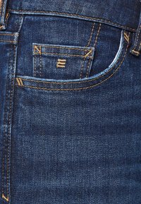 Los vaqueros de mezclilla azul oscuro presentan costuras amarillas en contraste, un pequeño bolsillo frontal y una textura suave sin adornos visibles.
