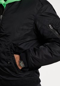 Svart bomberjacka med en slät, glänsande tyg. Har ribbade ärmslut och midja, en dragkedjeficka på ärmen och dolda sidfickor.