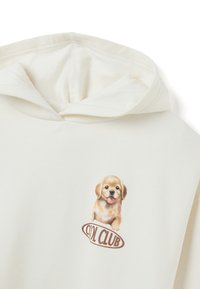 Crème-kleurige hoodie met een cartoon puppy graphic met een glimlach en de tekst "COOL CLUB" in bruin eronder. Zachte stof structuur.