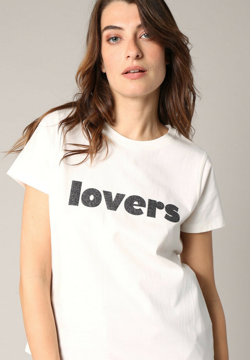 Witte katoenen T-shirt met een ronde hals en korte mouwen. Op de voorkant staat het woord "lovers" gedrukt in vetgedrukte, zwarte glanzende letters.