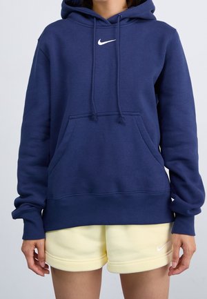 Person iført en marineblå Nike hættetrøje og lysegule Nike shorts, stående mod en ensfarvet lys baggrund.