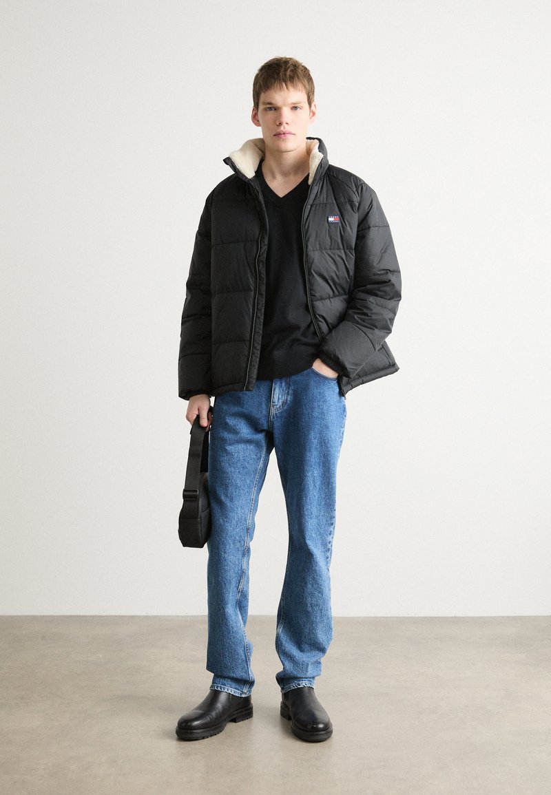 Schwarze Pufferjacke mit einem cremefarbenen Kragen, schwarzes T-Shirt, blaue Jeans und schwarze Lederstiefel. Das Modell hält eine schwarze Umhängetasche.