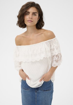 Žena s krátkymi tmavými vlasmi, oblečená v bielej čipkovej blúzke s odhalenými ramenami a modrej rifľovej sukni, ruky v vreckách, čelí dopredu.