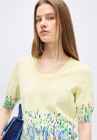 Νεαρή γυναίκα με ίσια ανοιχτοκαφέ μαλλιά φοράει απαλό κίτρινο πουλόβερ με πολύχρωμο αφηρημένο μοτίβο στα μανίκια και στο τελείωμα, κρατώντας μπλε τσάντα.