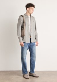 Grijze overhemd, wit t-shirt, blauwe jeans, bruine schoudertas, grijze schoenen, staande op een neutrale achtergrond.
