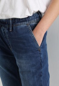 Pantalones vaqueros en azul oscuro con cintura elástica y bolsillos laterales. La tela tiene una textura desgastada y detalles con costuras.