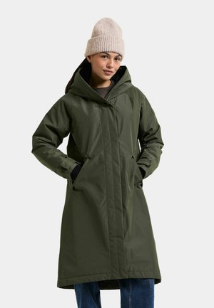 Kvinna klädd i en lång olivgrön huva, beige stickad mössa och blå jeans, ståendes med händerna i rockfickorna.