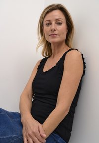 Černý žebrovaný top na ramínka s vlnitým výstřihem. Modelka má dlouhé rovné blond vlasy a sedí opřená o bílou zeď, na sobě má modré džíny.