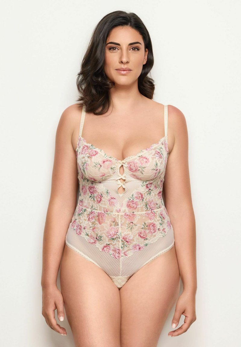 Nainen, jolla on keskipitkä tumma hius, pukeutuneena beigeen kukkakuvioiseen pitsibodysuitiin, jossa on vaaleanpunaisia ruusuja, seisoo yksinkertaista vaaleaa taustaa vasten.