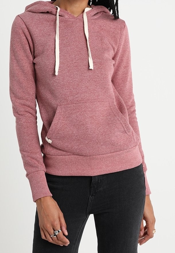 Femme portant un sweat à capuche rose poussiéreux avec des cordons de serrage blancs et un jean noir, debout devant un fond blanc uni.