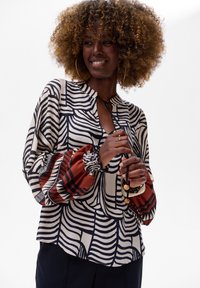 Bluzka w wzory z mieszanką czarnych i białych pasków oraz ziemistoczerwonymi akcentami, okrągły dekolt i bufiaste rękawy z elastycznymi mankietami.