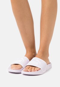 Világos lila csúsztatós papucsok, textúrázott pánttal, amelyen fehér Puma logo található. Nyitott orrú design, párnázott talppal a kényelem érdekében.