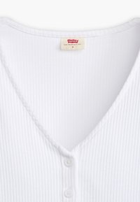 Valkoinen ribattu kangastoppi, jossa on V-mallinen kaula-aukko, ja etuosassa kolme napin kiinnitystä. Tunnuksessa lukee "Levi's" ja koko S.