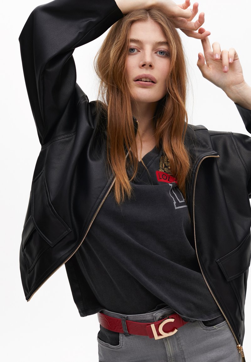 Černá bomber bunda z umělé kůže s oversized rukávy, kombinovaná se šedým tričkem s červeným vyšitým logem a červeným lesklým páskem.