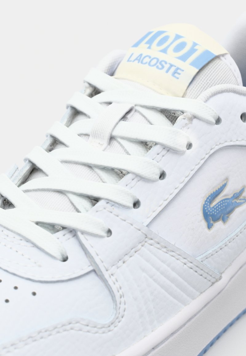 Witte leren sneaker met blauwe accenten. Beschikt over platte witte veters, geperforeerde neus en een blauw Lacoste-logo aan de zijkant.