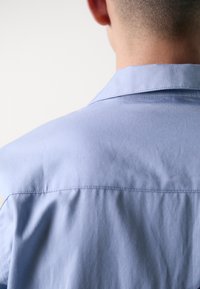 Camicia azzurro chiaro con bottoni, caratterizzata da una texture morbida e liscia, colletto dritto e pieghe sottili sulle spalle.
