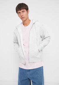Gray zip-up hoodie with kangaroo pockets, worn over a light pink shirt and blue denim pants. Smooth fabric, casual fit.