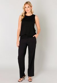 Schwarzes ärmelloses Oberteil kombiniert mit schwarzen Hosen. Lockerer Schnitt, weicher Stoff und gerades Bein. Mit Seitentaschen und offenen Absatzschuhen.
