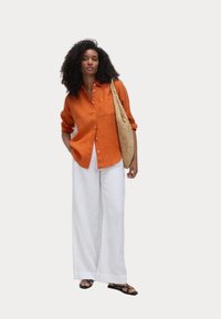 Rood-oranje linnen button-up shirt, witte wijde broek, gevlochten tan tas en zwarte sandalen. Het shirt heeft een borstzak en opgerolde mouwen.