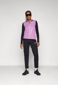 Gilet stretch violet avec poches zippées, associé à un haut à manches longues noir et un pantalon noir. Le modèle porte des lunettes de soleil et des baskets noires.