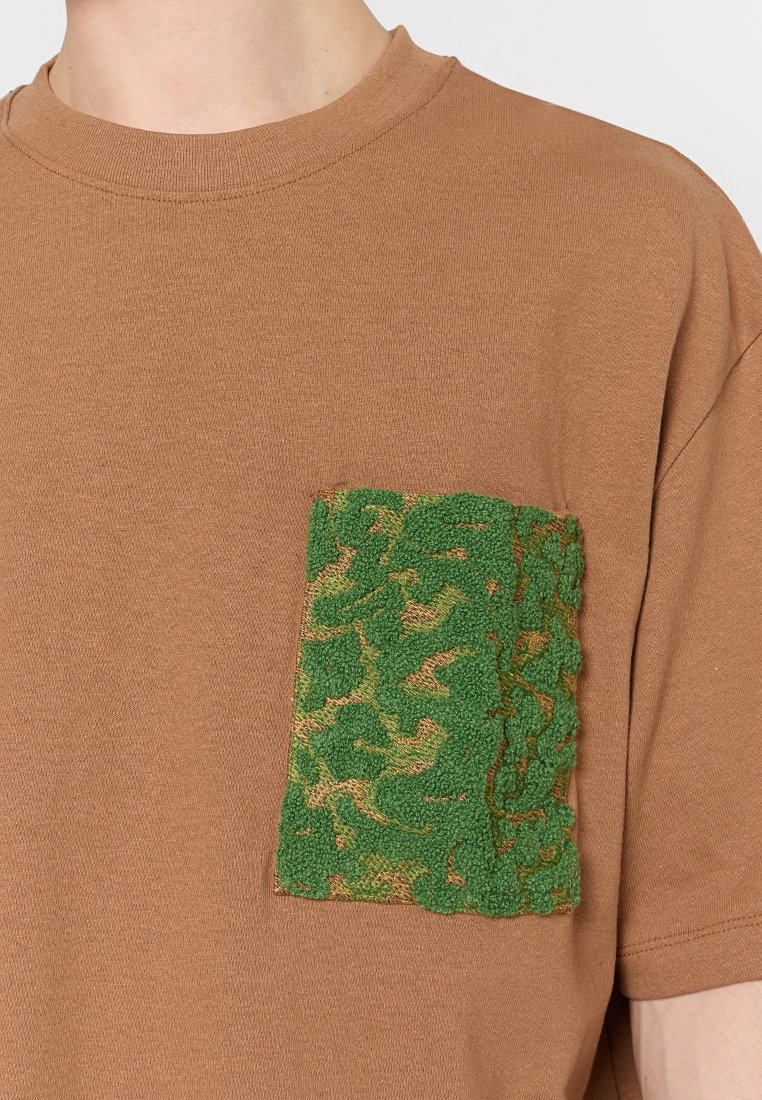 Brązowa koszula z krótkim rękawem z zieloną teksturowaną prostokątną kieszenią na lewym czole.