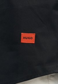 Μαύρο ύφασμα με πορτοκαλί ορθογώνιο ταμπέλα που φέρει το όνομα της μάρκας "HUGO" με μαύρα έντονα γράμματα.