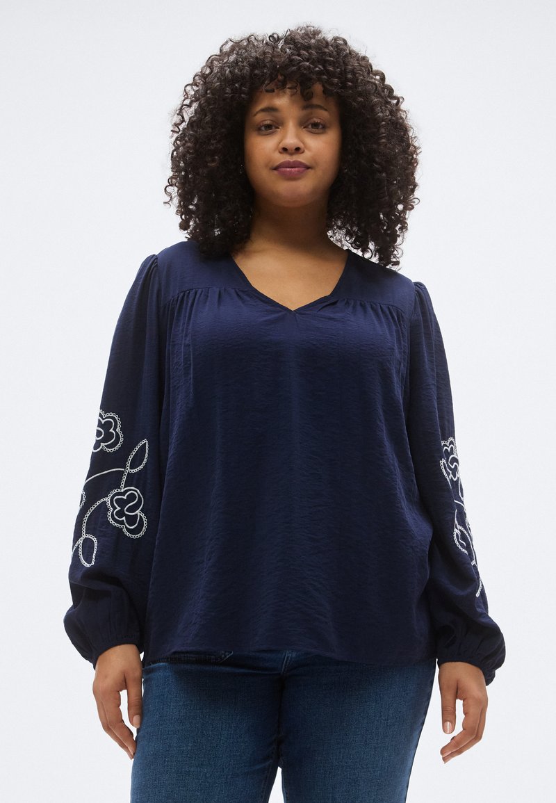 Blouse bleu marine avec un col en V, manches longues bouffantes et broderies florales blanches sur les manches. Confectionnée dans un tissu léger et texturé.